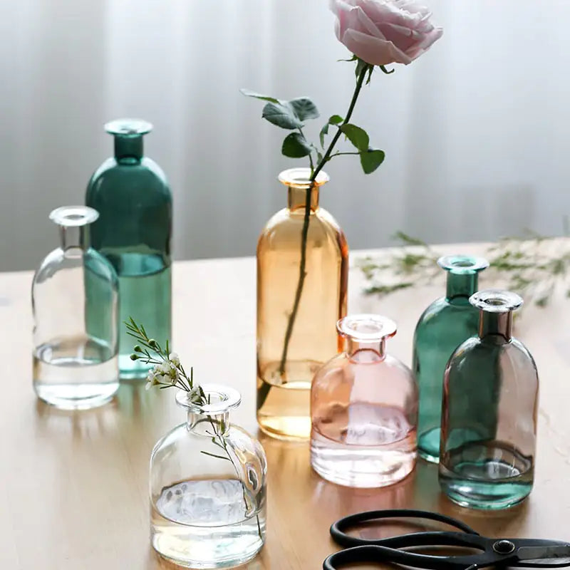Assorted colored glass vases with flowers on a wooden table in soft natural light